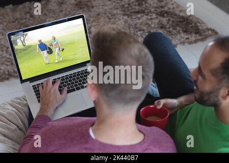 Two diverse male friends at home watching senior couple playing golf players on laptop. sports, competition, entertainment and technology concept digi Stock Photo
