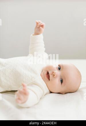 Smiling baby boy (2-5 months) in white outfit lying on bed Stock Photo