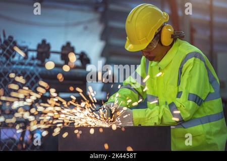 A worker is using a steel grinder in the factory. Industrial Worker Using Angle Grinder and Cutting a Metal Tube. Worker in Safety Uniform and Hard Ha Stock Photo