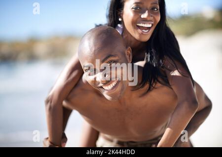 My man is so strong. A handsome african-american man giving his girlfriend a piggyback on the beach. Stock Photo