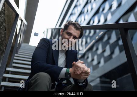 young male business analyst in business clothes sits smiling on the stairs of the business center Stock Photo
