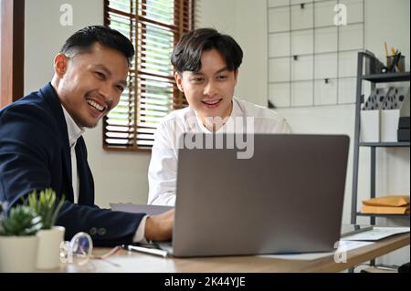 Cheerful and smart Asian businessman working with his coworker in the office. Male marketing employee shows his project plan on the laptop screen to h Stock Photo