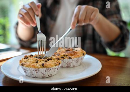 Young Asian female enjoys eating wholewheat muffin with raisins in the coffee shop, using fork and knife. cropped and close-up image Stock Photo