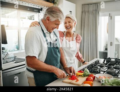 Senior couple, cooking and having fun while preparing a healthy food with vegetables for a vegan meal in the kitchen at home while laughing and having Stock Photo