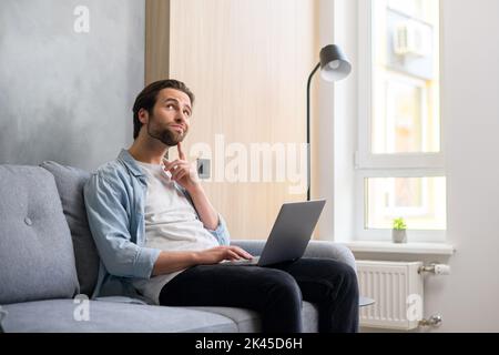 Pensive young businessman in casual wear sitting at the sofa at home, using laptop computer, watch away thoughtfully, looking for business tasks solving. Male employee on the couch Stock Photo