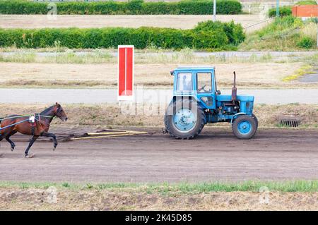 An old tractor prepares a route for horses on the racetrack of the hippodrome by leveling it with a tow hitch. Copy space. Stock Photo