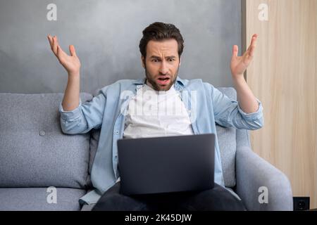 Young hipster guy sitting at the sofa in front of laptop and raising his hands up, feeling frustrated and scared from occurred issues. Businessman is upset to hear the unexpected bad news Stock Photo