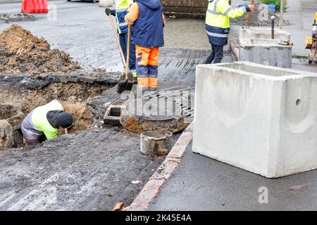 A group of road workers repair sewer storm hatches and change old concrete manholes for new ones on a roadway on an autumn day. Stock Photo