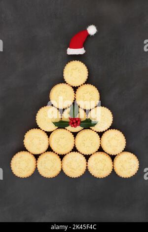 Festive Christmas tree concept shape with mince pies, santa hat, winter holly with red berries on grunge grey background. Abstract surreal food design