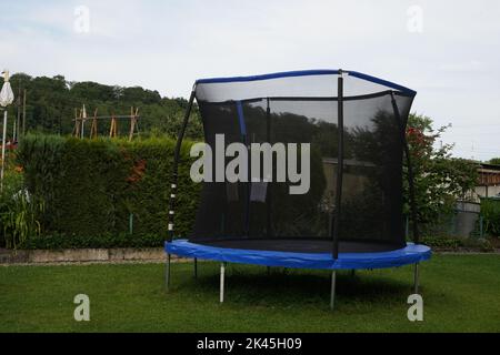 Empty blue trampoline with safety net on the lawn in garden. In the background is green vegetation creating hedge. Stock Photo