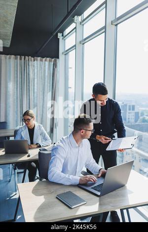 Group of young business colleagues in classic clothes sitting at table with laptop and documents, discussing details, looking at each other. The atmos Stock Photo