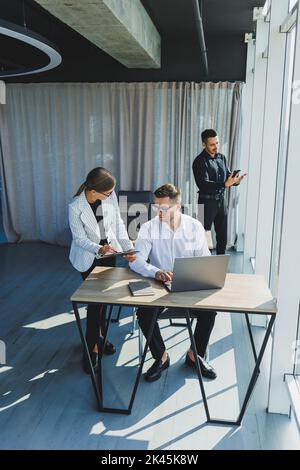 Group of young business colleagues in classic clothes sitting at table with laptop and documents, discussing details, looking at each other. The atmos Stock Photo