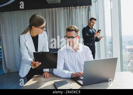Group of young business colleagues in classic clothes sitting at table with laptop and documents, discussing details, looking at each other. The atmos Stock Photo