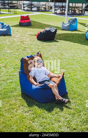 A fair-haired boy of 5-6 years old in a white T-shirt and shorts is sitting on a bean bag chair. A child in sunglasses rests in the fresh air Stock Photo