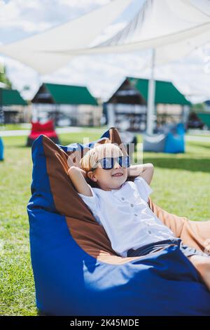 A fair-haired boy of 5-6 years old in a white T-shirt and shorts is sitting on a bean bag chair. A child in sunglasses rests in the fresh air Stock Photo