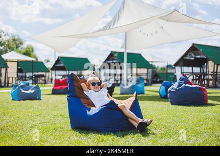 A fair-haired boy of 5-6 years old in a white T-shirt and shorts is sitting on a bean bag chair. A child in sunglasses rests in the fresh air Stock Photo