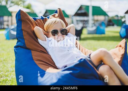 A fair-haired boy of 5-6 years old in a white T-shirt and shorts is sitting on a bean bag chair. A child in sunglasses rests in the fresh air Stock Photo