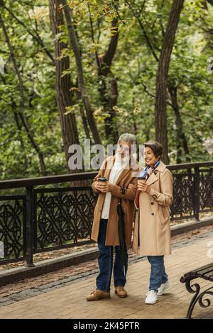 full length of cheerful senior couple in beige coats holding paper cups and walking in park,stock image Stock Photo