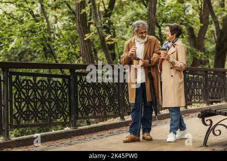 full length of cheerful retired couple in beige coats holding paper cups and walking in park,stock image Stock Photo