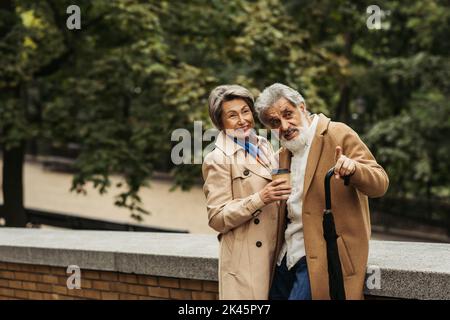 senior man holding umbrella and pointing with finger near cheerful wife with paper cup,stock image Stock Photo
