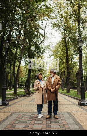 full length of happy senior woman holding paper cup while walking with stylish husband in coat,stock image Stock Photo