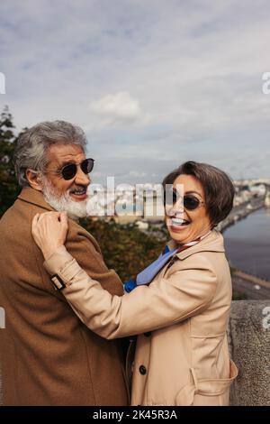 excited senior woman in stylish sunglasses hugging bearded husband in beige coat,stock image Stock Photo