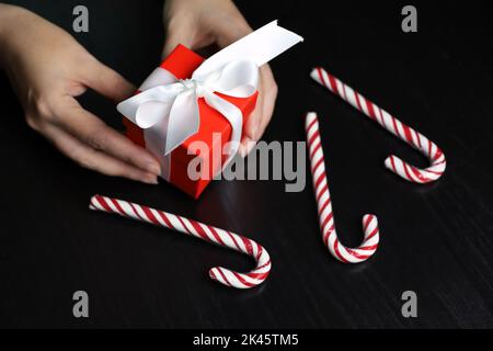 Red gift box in female hands on dark wooden table with candy canes. Concept of Christmas holiday, romantic surprise Stock Photo