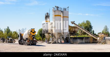 A concrete mixer truck and a wheel loader are parked next to sand silos in a quarry on a sunny day. Stock Photo