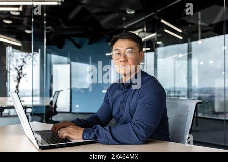 Serious and focused businessman looking at camera, asian man working inside modern building, man wearing glasses and casual shirt using laptop. Stock Photo