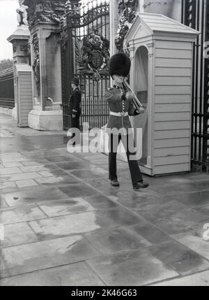 1958, historical, a policeman and a uniformed Queen's guard on duty at the entrance outside Buckingham Palace, Westminster, London, England, UK. Guardsman in their tunics and bearskins have been on sentry duty outside Buckingham Palace, the Queen's official residence, since Queen Victoria moved ther ein 1837. Stock Photo