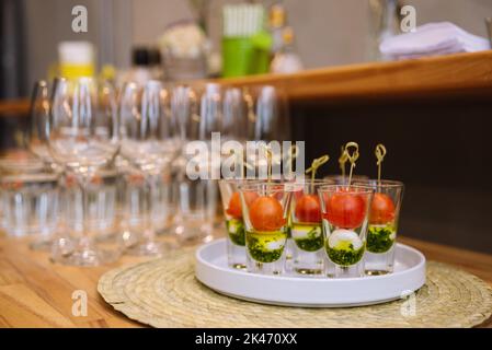 Caprese canapés in shot glasses on a skewer. cherry tomatoes, mozzarella cheese balls, pesto sauce. table with appetizers and empty wine glasses in th Stock Photo