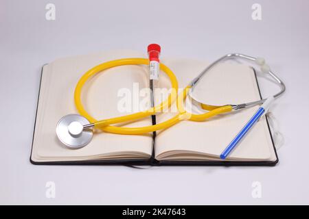 A medical stethoscope with a yellow heart on an open notebook with a blood tube, and a blue pen for the medical concept Stock Photo