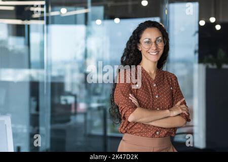 Portrait of a young beautiful Hispanic woman. Curly Latin American student in glasses stands in the hall of the university, college, looks at the camera, smiles. Stock Photo