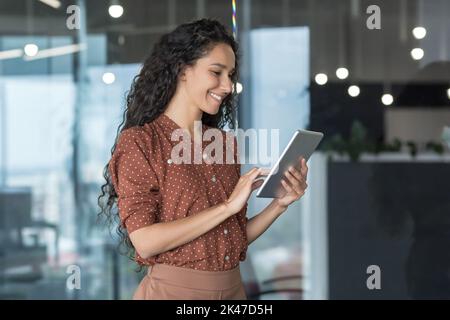 Young beautiful hispanic businesswoman standing in the office in the corridor. He holds a tablet in his hands, uses it, types, smiles. Stock Photo