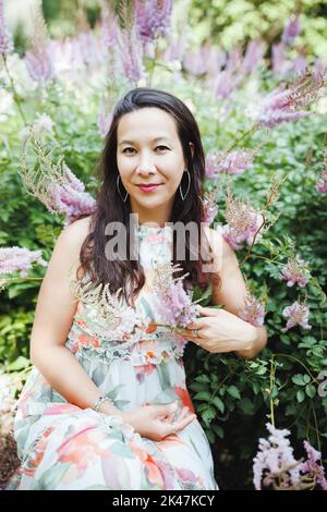 Beautiful happy mixed race Japanese woman in romantic floral maxi dress in summer park, smiling. Close-up portrait. Sunlight, copy space Stock Photo