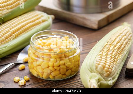 Pickled sweet corn in a glass jar, fresh corncobs, saucepan on kitchen table. Stock Photo