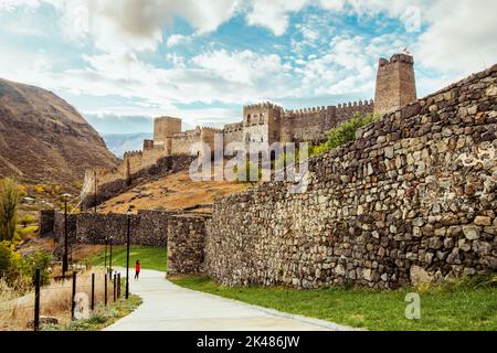 Female tourist stands on the path next to huge Khertvisi fortress wall with towers above in cloudy day. Travel copyspace background. Stock Photo