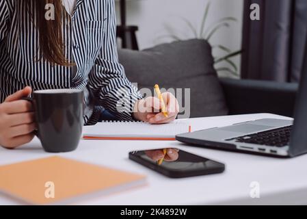 Close up of female hands holding cup coffee and pen making notes in diary sit at desk with laptop. Businesswoman writing business letter at work. Free Stock Photo