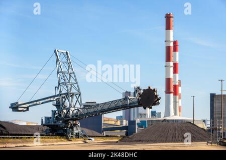 A bucket-wheel stacker-reclaimer and coal stockpiles in a thermal power station with red and white smokestacks on a sunny day. Stock Photo