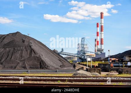 Coal stockpiles and bucket-wheel stacker-reclaimer in a coal-fired power station with red and white smokestacks on a sunny day. Stock Photo