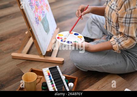 Cropped image of young female artist holding palette and painting picture on canvas with oil paints in home studio Stock Photo