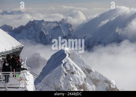 Besucher auf der Aussichtsplattform auf der Zugspitze genießen den eindrucksvollen Ausblick auf die verschneite Bergwelt der Alpen. Einige Gipfel sind von spannenden Wolken umhüllt. Unten links erkennt man die Tragseile einer Gondelbahn. Zaun und Dach sind verschneit. Stock Photo