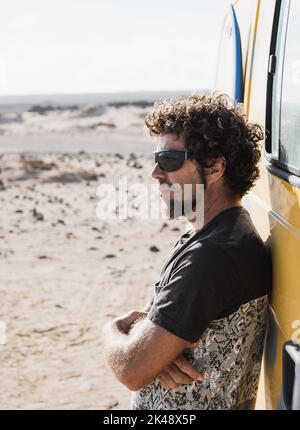 Caucasian man watching the landscape leaning on his camper van. He has a beard and he is wearing sunglasses Stock Photo