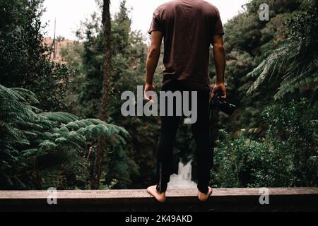 Barefoot caucasian boy in brown t-shirt and black pants standing on the wooden railing of a bridge with camera in hand looking at forest waterfall in Stock Photo