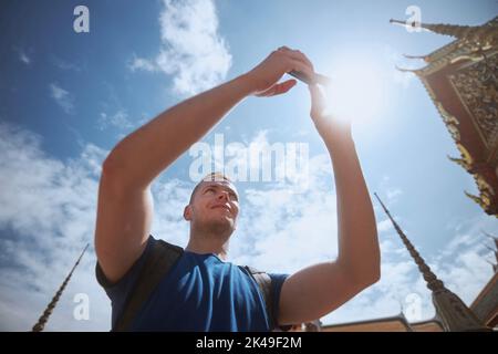 Man while taking pictures by mobile phone against roofs of landmarks. Tourist in Bangkok, Thailand. Stock Photo