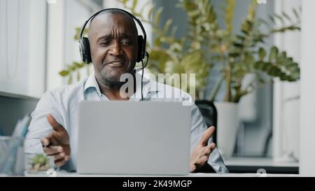 Senior african instructor teacher wears headphones using web camera for communication with clients teaching online video consultation during Stock Photo