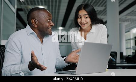 Arabian female leader instructor help african man colleague with computer work inspect trainee check mistakes on laptop. Two business people discuss Stock Photo