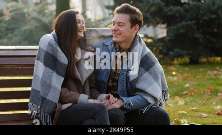 Cute loving couple sit on bench warming each other covering wool blanket outdoors in cold froze autumnal weather feel care love hugging funny talking Stock Photo