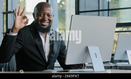 Happy african american businessman adult worker typing on laptop working remotely turn to camera smiling make approval good sign well done excellent Stock Photo
