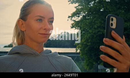 Fit sportswoman in grey hooded sportswear talking in virtual video chat calling by smartphone on street during morning sports training outdoors Stock Photo